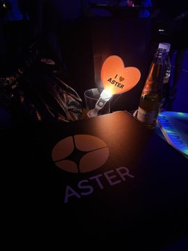 A dimly lit photo of a black shopping bag with the 'ASTER' logo and a glowing heart-shaped sign reading 'I ❤️ ASTER' next to a bottle in an ice bucket.