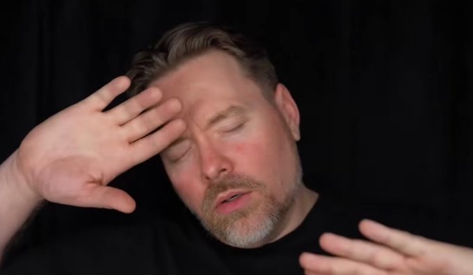 Photo of a man with closed eyes holding his hand to his forehead against a black background.