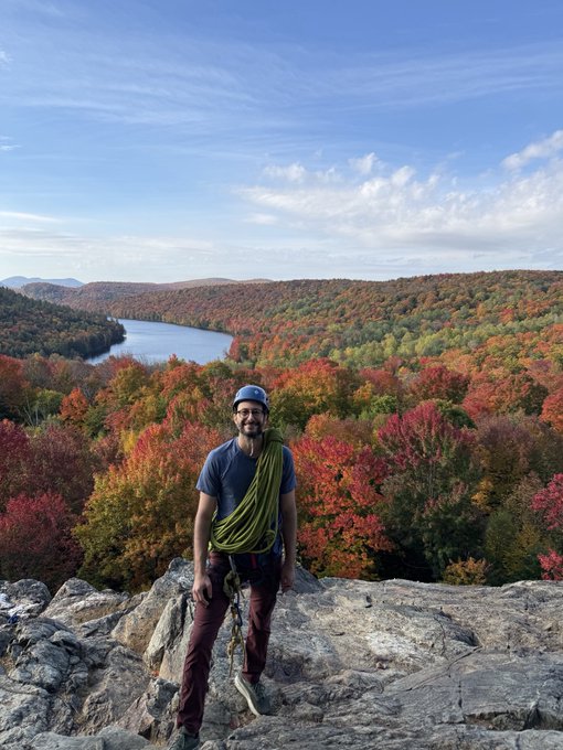 A rock climber smiles at the summit, draped in a coiled green rope, with a stunning panoramic view of Quebec's vibrant fall foliage and a winding lake behind him—a striking contrast to the tropical Miami he now calls home.