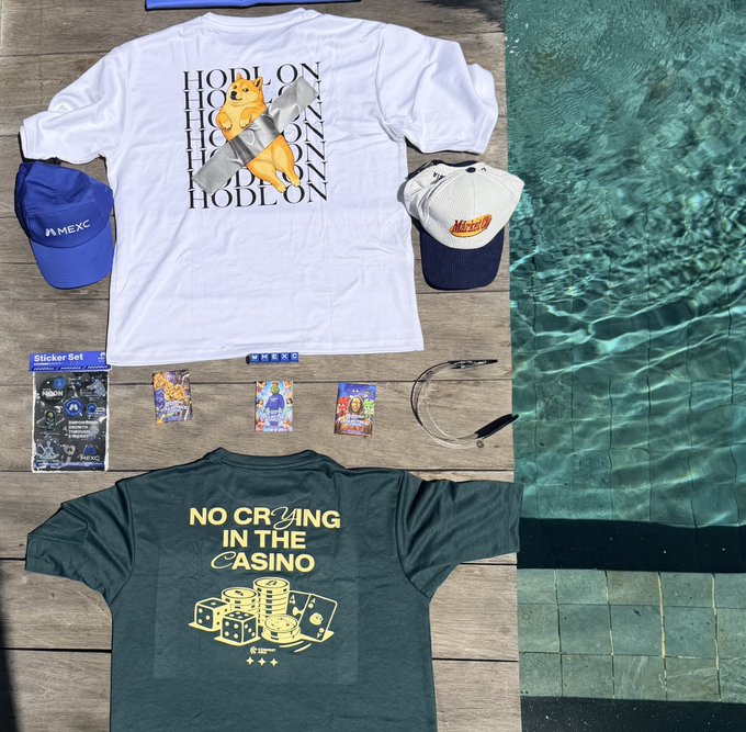 A flat lay of merchandise by a pool, including two MEXC exchange t-shirts—one white reading 'HODL ON' with a Shiba Inu dog and another green reading 'NO CRYING IN THE CASINO'—along with MEXC branded hats, stickers, and lanyards.