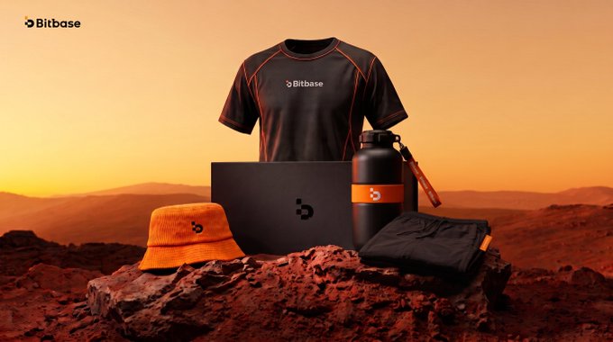 A Bitbase promotional product display featuring a black shirt, water bottle, laptop, hat, and bag arranged on rocks against an orange Martian-like landscape.