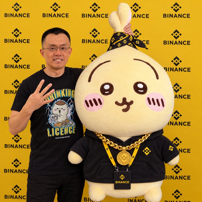 A man wearing a black 'THINKING LICENCE' t-shirt posing with a large plush mascot wearing a bandana and gold chain, with a Binance logo backdrop.