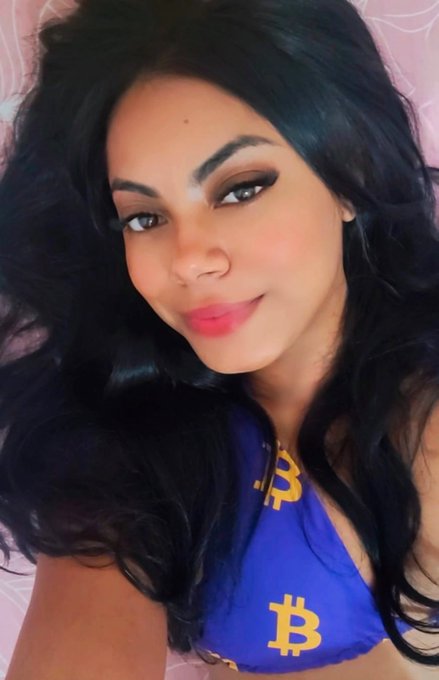 A selfie of a woman with long, voluminous black hair and pink lipstick, wearing a blue top featuring gold Bitcoin (₿) logos, likely promoting cryptocurrency airdrops tied to $LINA.