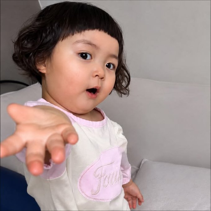 A young child with short curly hair wearing a cream and pink outfit with "Jou" embroidered on it reaches out toward the camera.