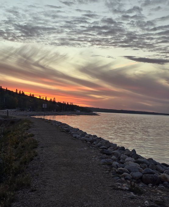 A sunset view over a large body of water with scattered clouds in the sky, featuring a gravel shoreline lined with rocks and silhouetted trees in the distance.
