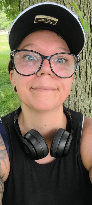 A close-up selfie of a person wearing glasses, a black "ASSOCIATION" baseball cap, and black Beats headphones around their neck, standing in front of a tree.