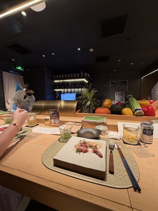 A lively dinner with @loldefi and @jimtalbot at a modern restaurant, where a beautifully plated dish sits beneath a colorful display of fruit and vegetables. A can labeled “SOLO” is visible on the table, matching the post’s playful “Whimsy maxing” mood.
