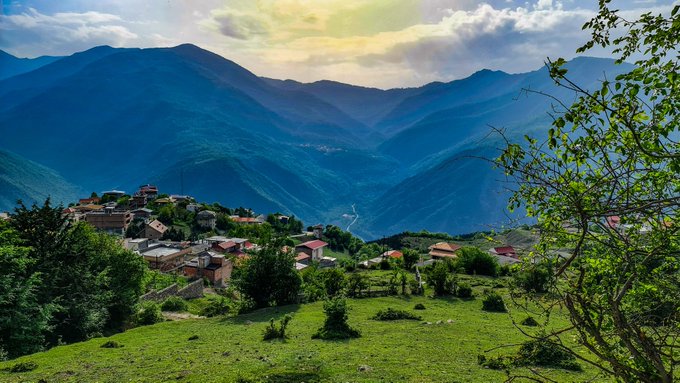 A scenic landscape featuring a small village nestled in lush green mountains under a cloudy sky with sunlight breaking through.