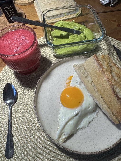 A breakfast plate featuring a sunny-side-up egg and bread, served alongside a glass of pink smoothie and a glass container of avocado or matcha spread.
