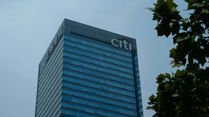 An upward view of a tall glass office building with the 'citi' logo and red arc at the top, framed by tree leaves against a pale sky.