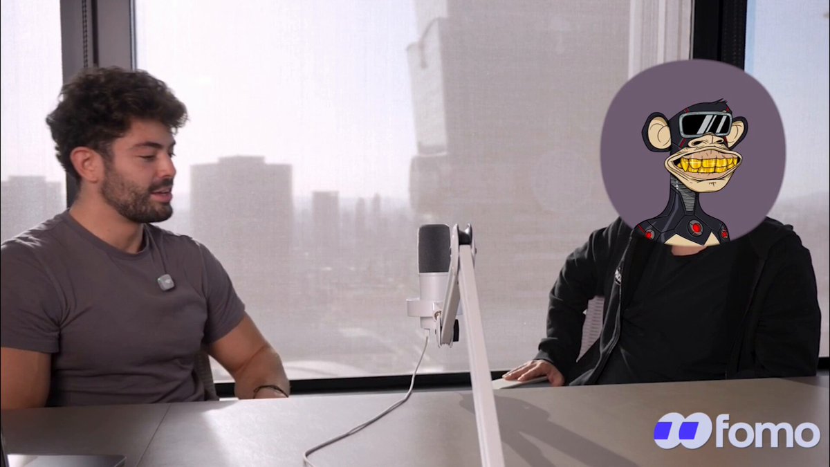 The image features two men sitting at a table with microphones set up between them. One of the men has a cartoon monkey head, which appears to be wearing sunglasses and a hat. The other man seems to be engaged in conversation or listening to something on the microphone. In the background, there is a window that provides natural light into the room.