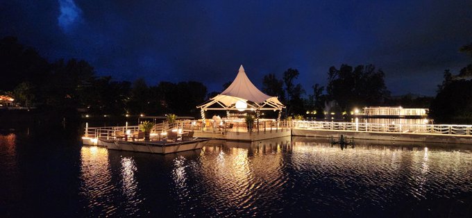 A nighttime photograph of an illuminated waterfront pavilion or restaurant with a peaked white tent-style roof, situated on a pier over a calm lake with lights reflecting on the water and dark trees in the background.
