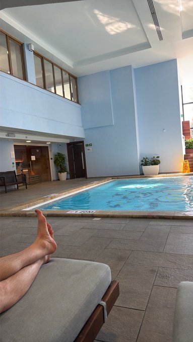 A pair of bare feet rest on a lounge chair by an indoor pool, with the swimmer's toes pointing toward the water in a relaxed pose, tying into the playful contest prompt asking followers to guess whose feet appear in the shot.