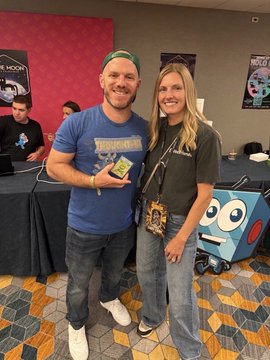 Photo of a man in a blue Jurassic Park t-shirt and a woman in a black t-shirt posing together at what appears to be a gaming or collectibles convention.