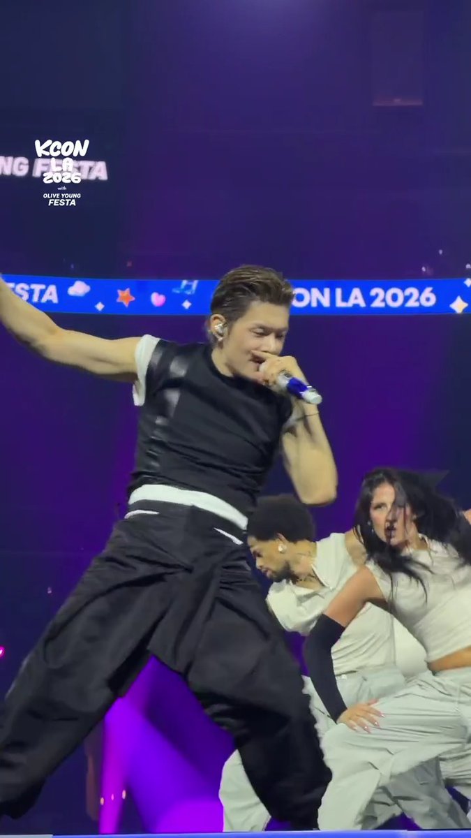 Yeonjun of TXT performs energetically on the KCON LA 2026 Counting Stage, gripping a microphone while dancing with backup dancers in white, with "KCON LA 2026" banners visible in the purple-lit background.