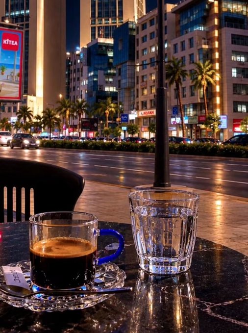 A nighttime view from an outdoor cafe table featuring a small glass cup of espresso and a glass of water, looking out onto a wet city street with illuminated storefronts, including a sign reading 'XTEA'.