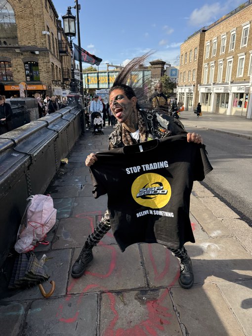 A person with punk styling holding up a black t-shirt that reads "STOP TRADING BELIVE IN SOMETHING" with a Yellow Sun SPX logo, standing on a city sidewalk with graffiti.