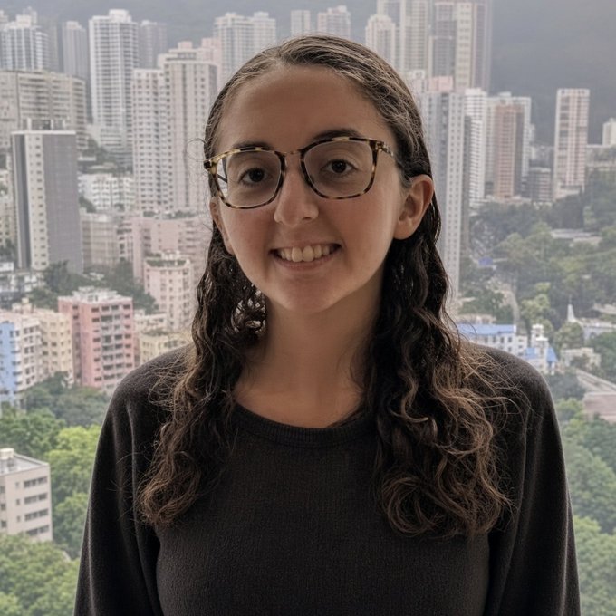 A young woman with curly brown hair and glasses smiles while wearing a dark sweater, standing in front of a window with a dense city skyline and mountains in the background.