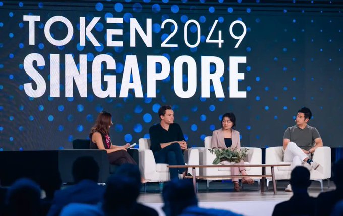 The image features a group of four people sitting around a table, with one person holding a microphone. They appear to be engaged in a discussion or presentation related to space exploration. The backdrop for this gathering includes a large blue sign that reads " Token 2049 Singapore", indicating an event or conference taking place in Singapore.