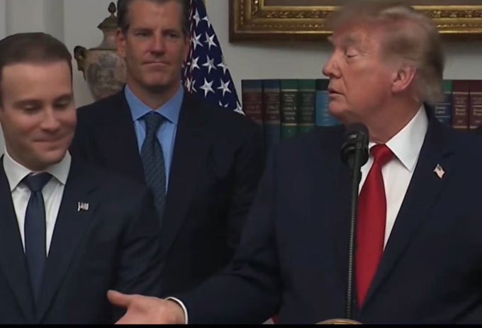 A photo of Donald Trump standing at a microphone speaking, with two other men in suits standing beside him in front of an American flag.