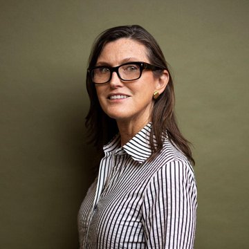 A woman with long brown hair and dark-rimmed glasses, wearing a black and white striped button-up shirt, smiles against an olive green background. The image does not show any visible text, numbers, or names, but it likely represents Cathie Wood, CEO of ARK Invest, whose recent $37 million Bitcoin purchase aligns with the post's message of continued institutional accumulation.