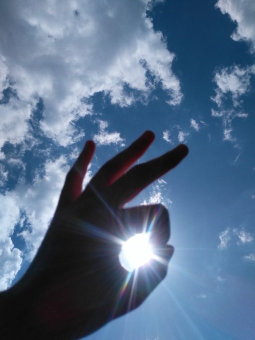 A silhouetted hand held up against a bright blue sky with white clouds, with the sun shining between the fingers.