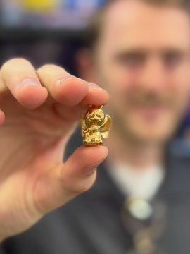 A man holding up a small golden figurine between his fingers, with his face blurred in the background in what appears to be an office or showroom setting.