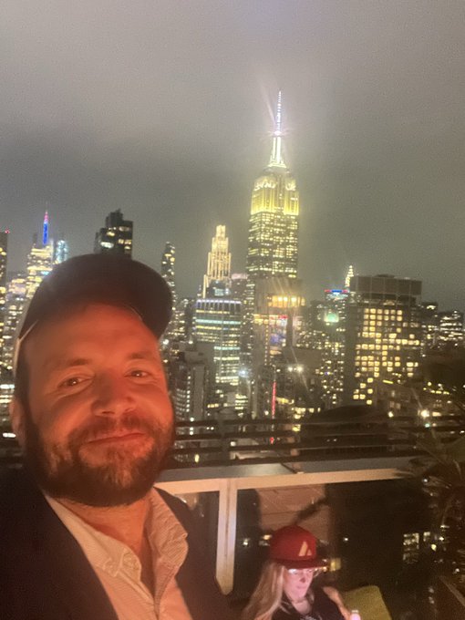 A selfie of a smiling bearded man wearing a baseball cap, taken at night with the illuminated New York City skyline, including the Empire State Building, in the background.