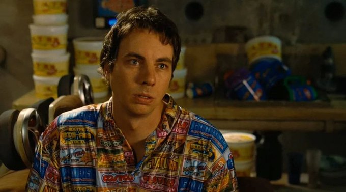A young man with a dazed, confused expression stares blankly forward while wearing a colorful shirt patterned with various snack food logos (including "OREO" and "Jell-O"), standing in what appears to be a cluttered storage room. The image humorously illustrates the post's point about people being "dumb and lazy" when it comes to crypto, with his vacant "derp" face serving as the perfect visual representation of someone unwilling to take self-custody responsibility for their own assets.