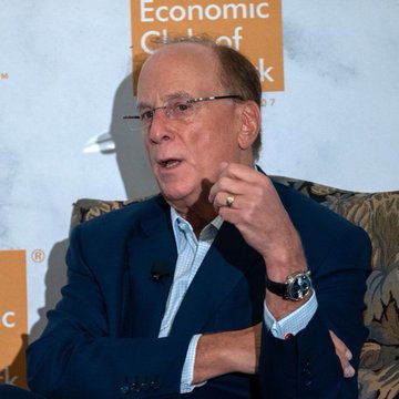 An older man with glasses wearing a navy blue blazer over a light shirt speaks at what appears to be the Economic Club, gesturing with his hand near his face.
