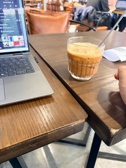 A glass of coffee with a spoon sits on a wooden cafe table next to an open laptop displaying a webpage.