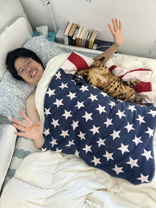 A smiling person with glasses lies in bed under a stars-and-stripes blanket, waving at the camera while a Bengal cat rests beside them.