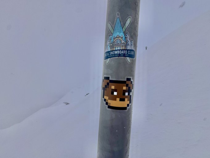 A pixel art sticker of a brown character with a bandolier is affixed to a metal pole bearing a 'Mt. Bachelor Ski & Snowboard Club' logo, set against a snowy background.