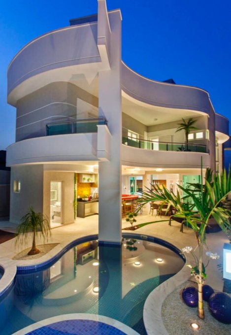 A luxurious multi-story home with modern curved architecture, featuring an open-concept living area, pool, and warm lighting at dusk.