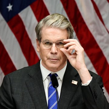Treasury Secretary Scott Bessent adjusts his glasses while speaking at a podium, with the American flag behind him. The image accompanies news that the U.S. is preparing to seize approximately $1 billion in Iran-linked cryptocurrency, with Bessent stating that the Treasury Department knows exactly where the assets are located.