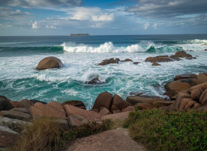A coastal landscape with crashing turquoise waves against rounded boulders under a cloudy blue sky.