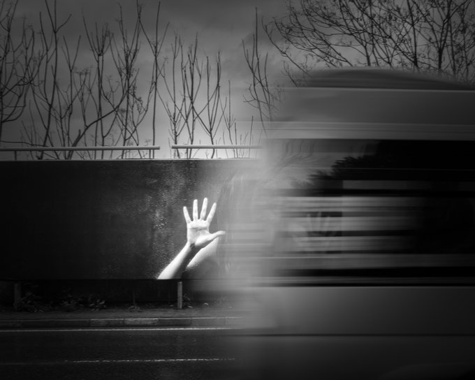 A moody black-and-white photograph showing a hand reaching up from behind a wall along a roadside, with a blurred vehicle speeding past, creating a haunting, ghostly atmosphere.