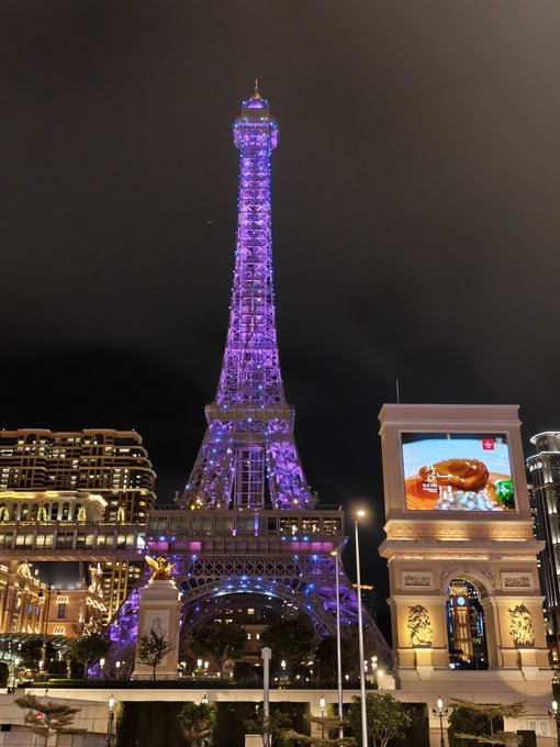 A replica of the Eiffel Tower illuminated in purple lights at night, with a digital billboard nearby and the Paris Las Vegas hotel in the background.