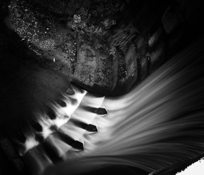A black-and-white long-exposure photograph captures water cascading over stone steps at the base of a waterfall, the silky flow of the current contrasting with the rough, rocky surroundings—evoking the "chasing the falls" theme of the artwork listed on Objkt.