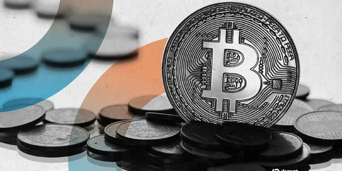 A physical Bitcoin token standing upright among a pile of silver coins, with a graphic overlay featuring teal and orange semicircles.