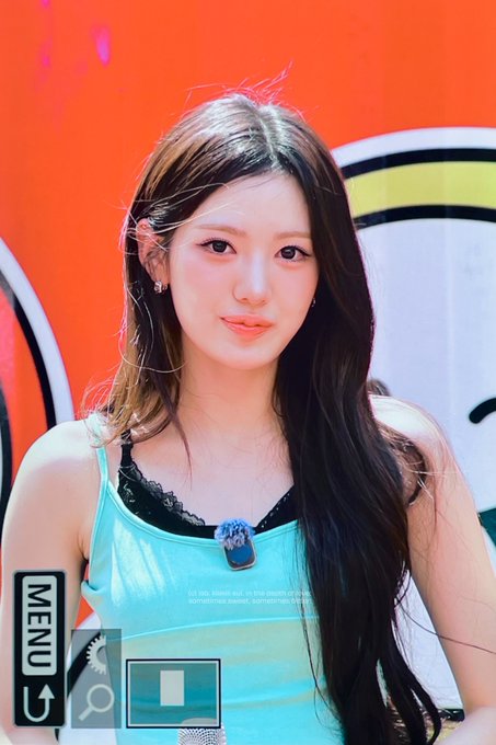A young woman with long dark hair wears a teal camisole with black lace trim and a small "KiiiKiii" pendant, posing in front of a vibrant orange backdrop. The image is a camera screen capture showing "MENU" and zoom icons, connecting to the post featuring SUI (수이) of the K-pop group KiiiKiii on their official account.