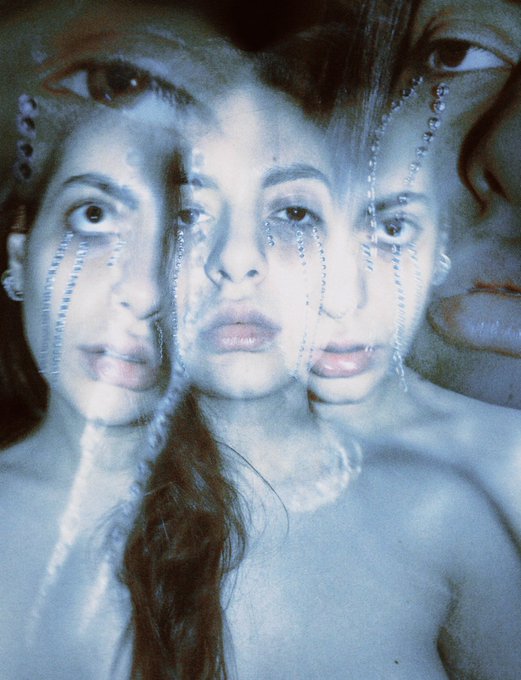 A haunting double exposure photograph of a young woman with beaded facial tears, layered with a close-up of another face, rendered in cool blue tones to evoke vulnerability and emotional conflict—mirroring the caption's plea to a figure who draws in broken people ("the martyr, the devil, the angel, the roadkill") and asks whether anything can convince them to stay.
