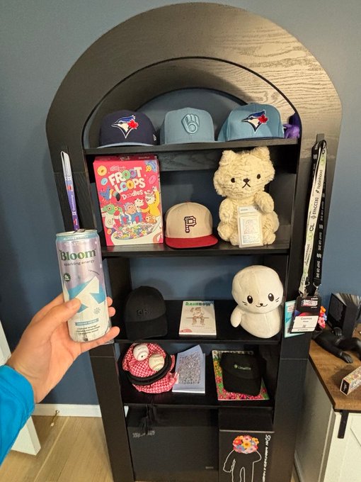 Photo of a hand holding a Bloom sparkling water can in front of a black arched display shelf containing various sports caps (Blue Jays, Brewers), a teddy bear, a plush toy, books, and lanyards.