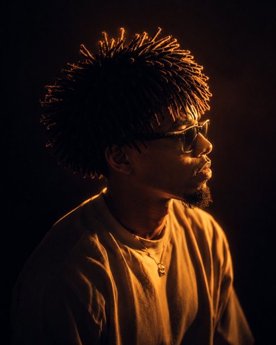 The image features a man with dreadlocks wearing glasses and sunglasses, who appears to be a hipster or artist. He has his head tilted back as if he's looking off into the distance. The background of the photo is dark, which contrasts with the bright light shining on him from above. There are no texts present in the image, but there is a caption that reads "gm to everyone It' s another week to grind and be productive. With the market condition right now, do you still think @quipnetwork will tge in Q2?".