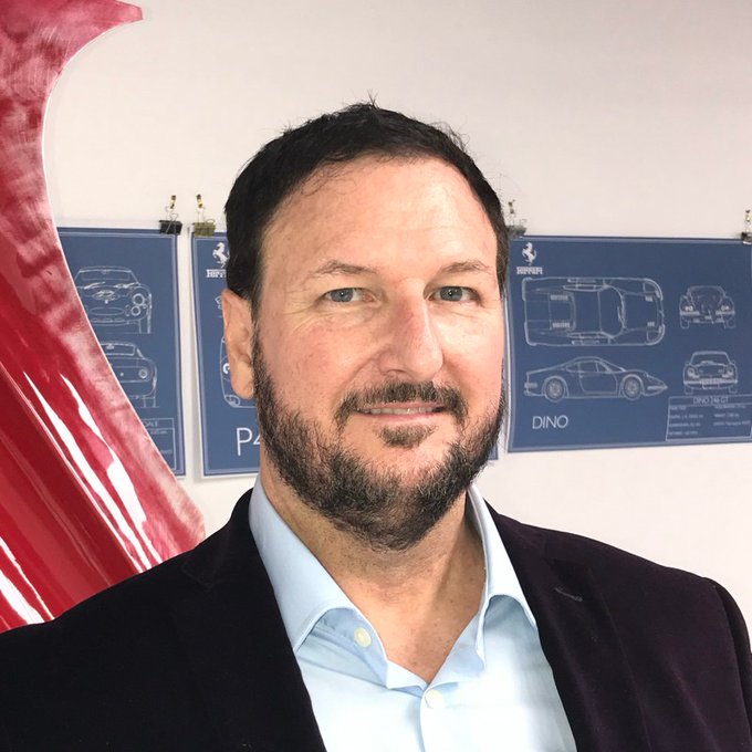 A selfie of a bearded man in a dark blazer and light blue shirt standing in front of blue Ferrari design blueprints labeled 'DINO' with a red Ferrari logo visible on the left.