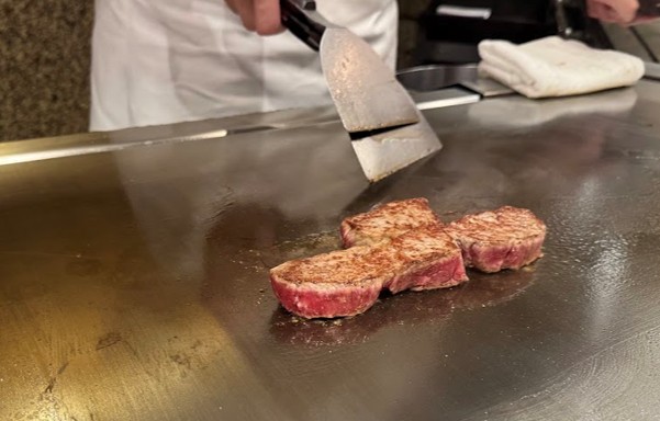 A chef uses a spatula to cook pieces of raw red meat on a large flat metal teppanyaki griddle.