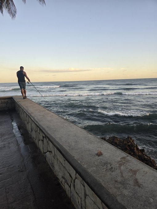 A person in dark clothing stands on a stone seawall at sunset, holding a fishing rod out over the ocean waves.