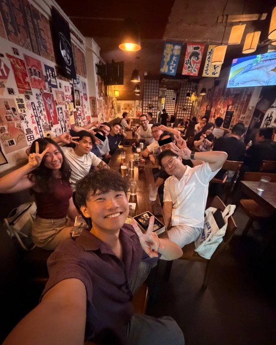 A selfie of a young man making a peace sign in a crowded, dimly lit restaurant or bar with Japanese-style wall decorations, surrounded by other patrons also posing.