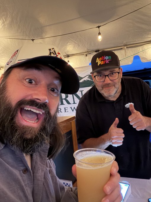 Two men pose enthusiastically at a brewery booth—one with a wide grin and full beard on the left, the other wearing an "Action Park" hat and giving two thumbs up on the right—while holding up a frosted cup of beer beneath a banner partially reading "RN" and "REW," capturing the celebratory moment of the brewery accepting Bitcoin via Square at the farm party.