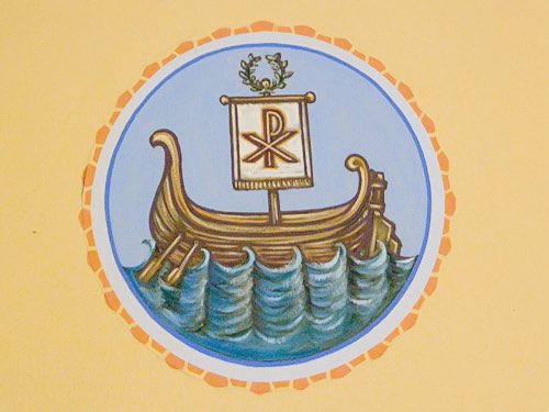 A circular illustration of a wooden ship sailing on blue waves, featuring a sail with a Chi-Rho symbol (☧) inside a blue and orange border.